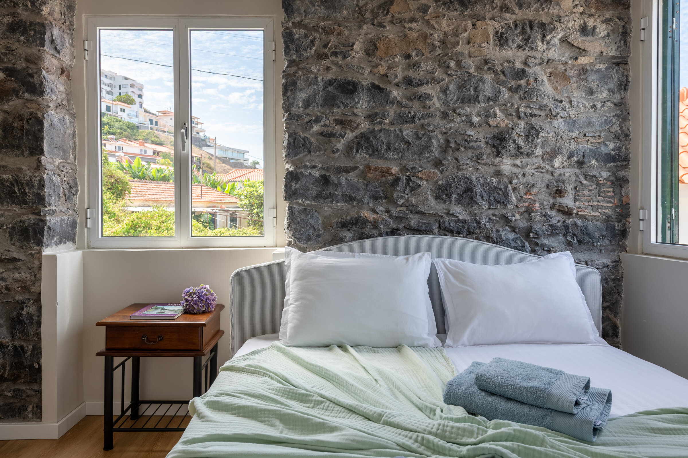 Bed with soft green linens against a stone wall, ocean-facing window